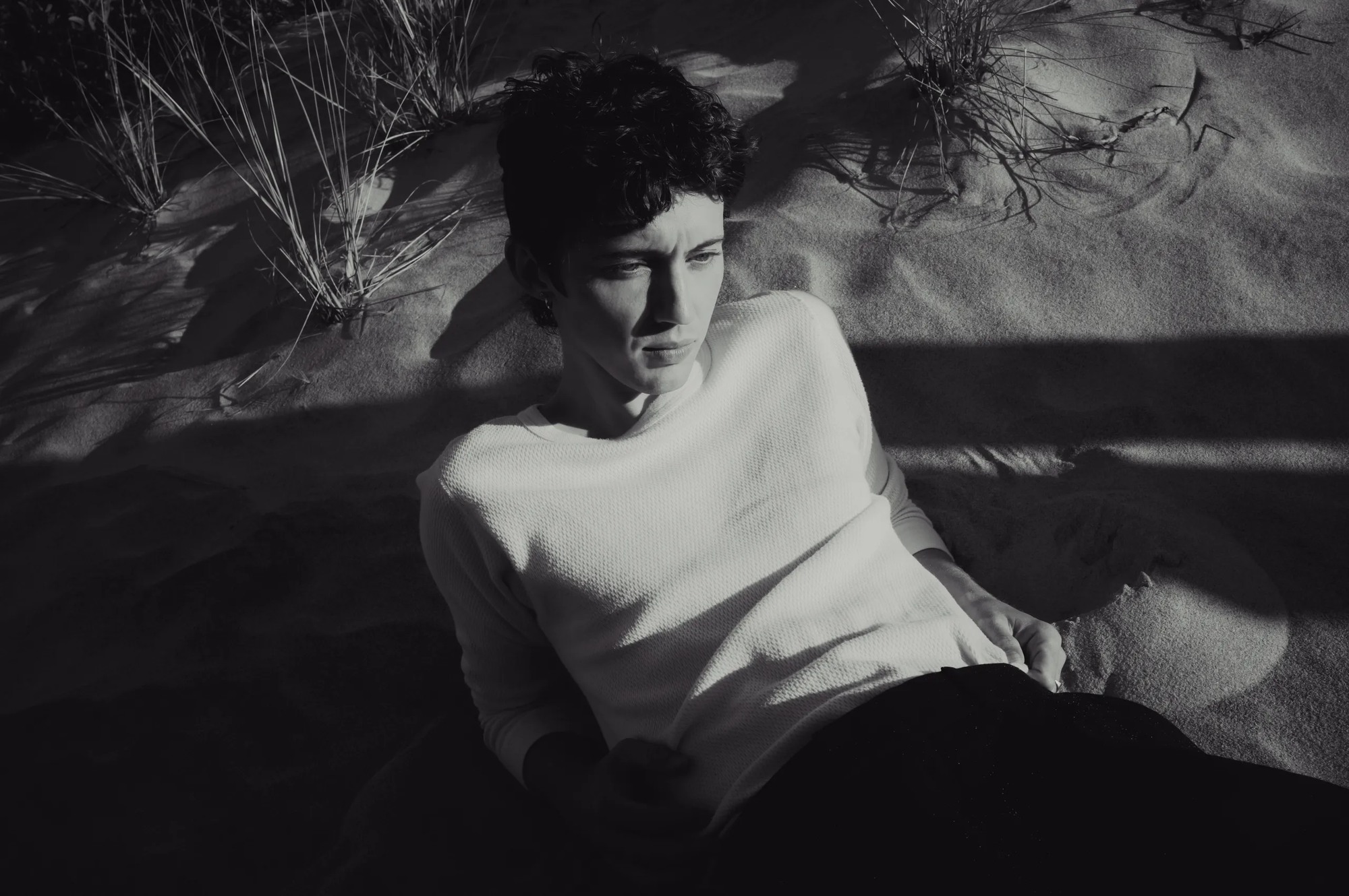 Troye Sivan deitado na areia, vestindo uma camisa branca, em fotografia em preto e branco